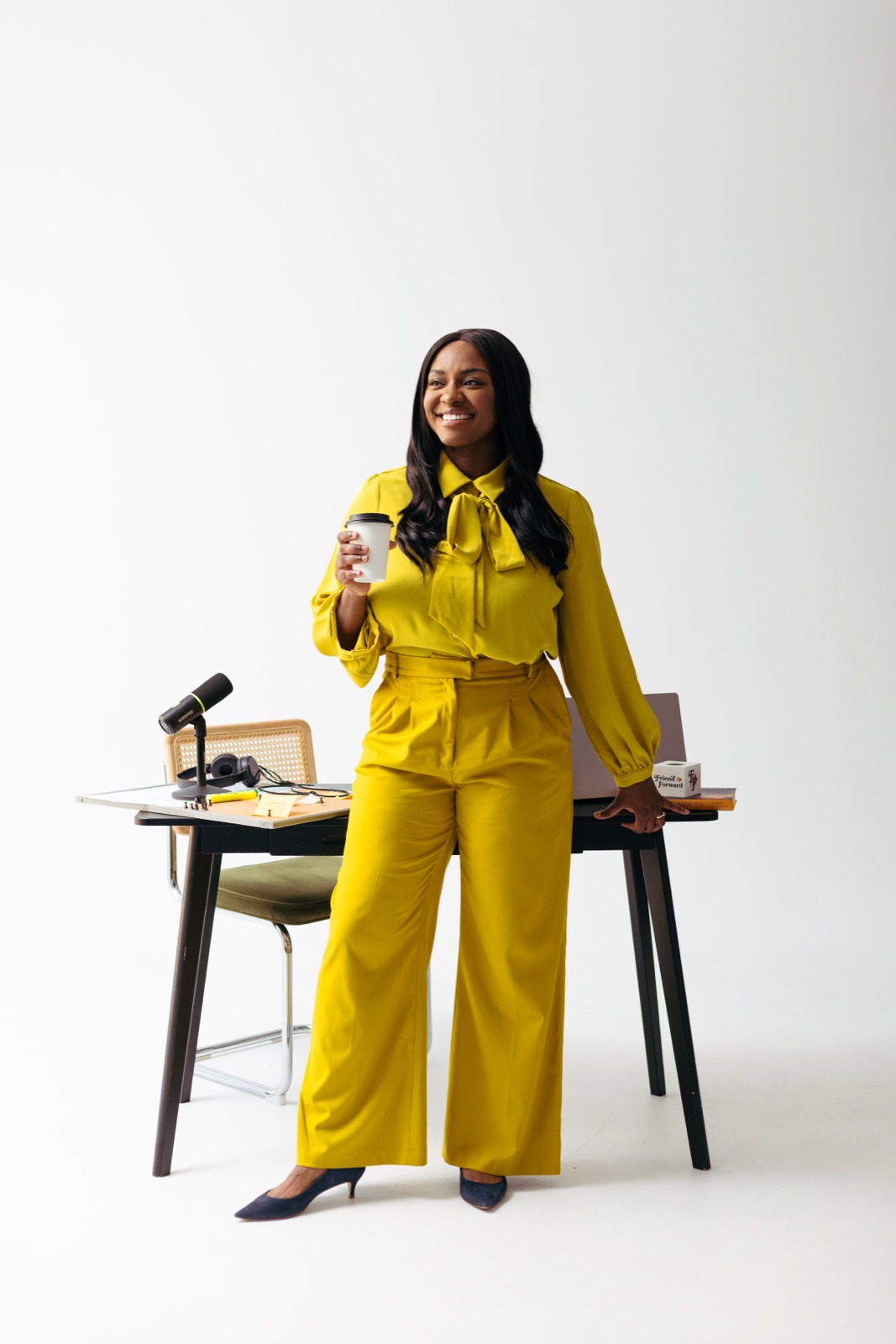 Danielle Bayard Jackson in a mustard suit, standing at a desk with a podcast microphone